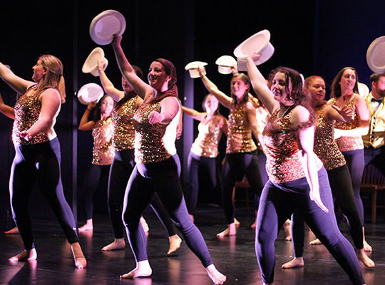 Dancers holding white hats and dressed in gold sequins fill the stage.