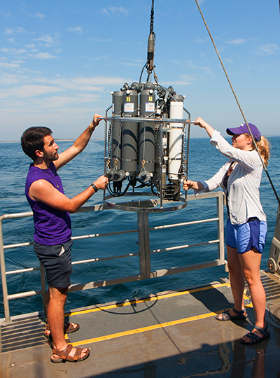 Students standing on the deck of a research vessel help lower canisters into the ocean water. 